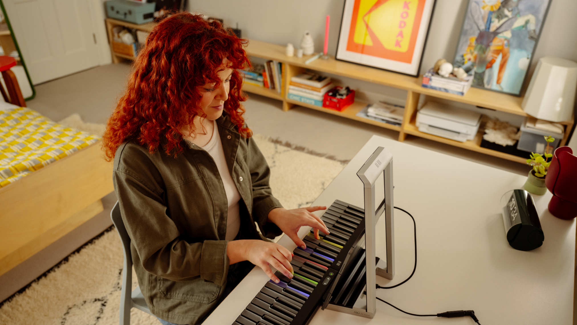 A person with curly red hair playing a ROLI Piano on a white desk in a cozy bedroom, with the instrument’s colorful illuminated keys and a ROLI Piano M stand visible.