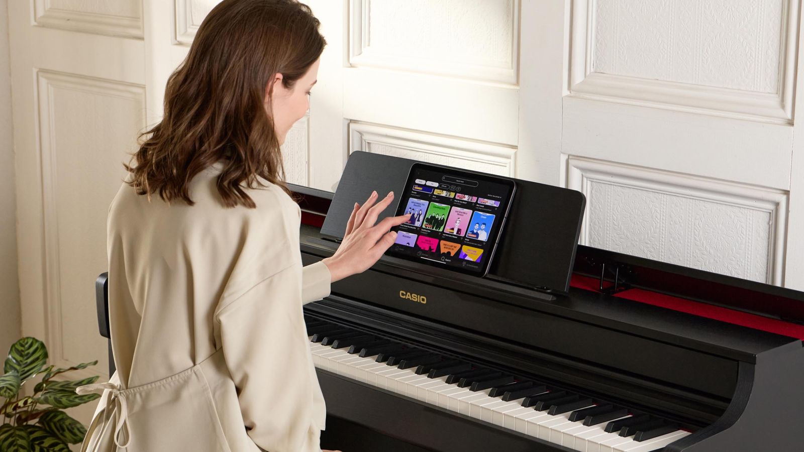 A woman seated at a black Casio digital piano uses a touchscreen tablet placed on the music stand, with colorful app icons visible on the screen in a home setting.