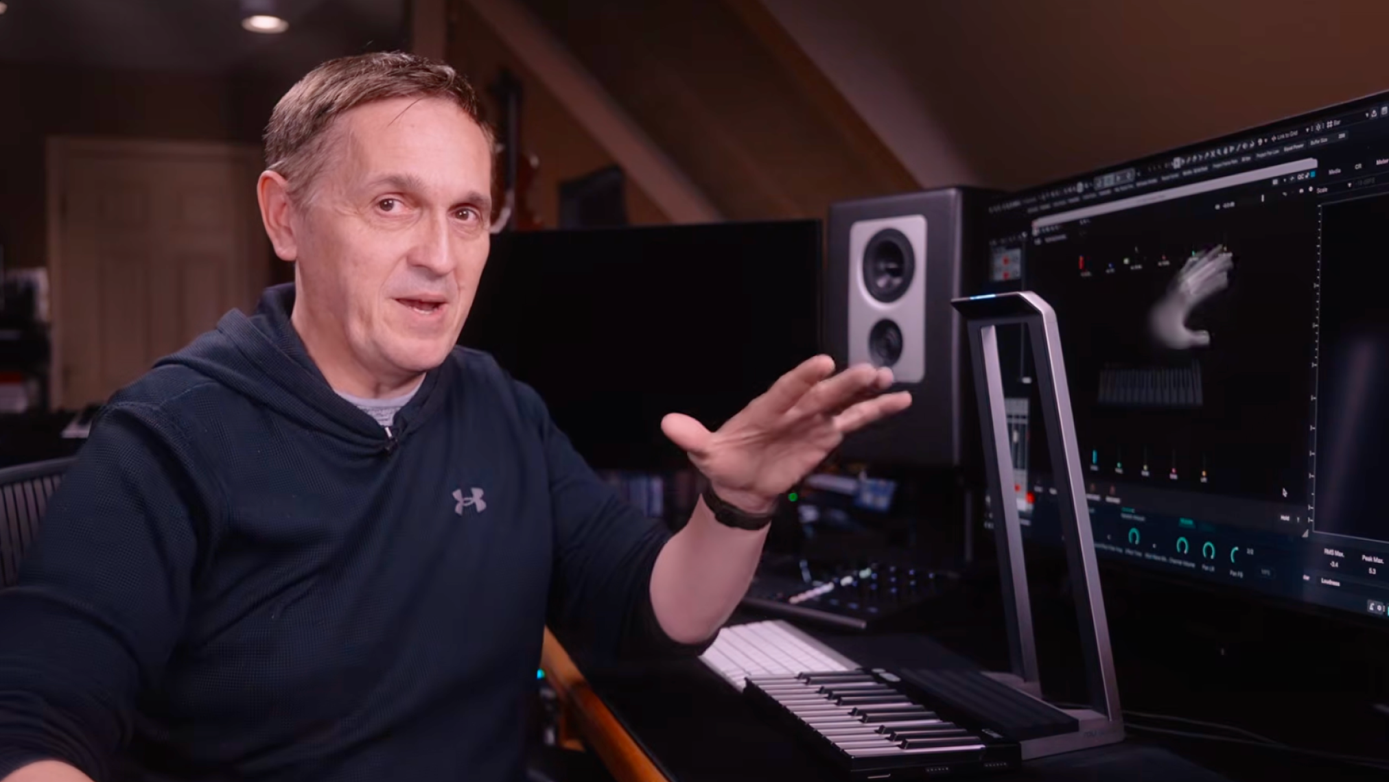A person demonstrating a ROLI Piano M and ROLI Airwave in a music studio, with their hand raised above the Airwave and a computer screen displaying hand tracking software.