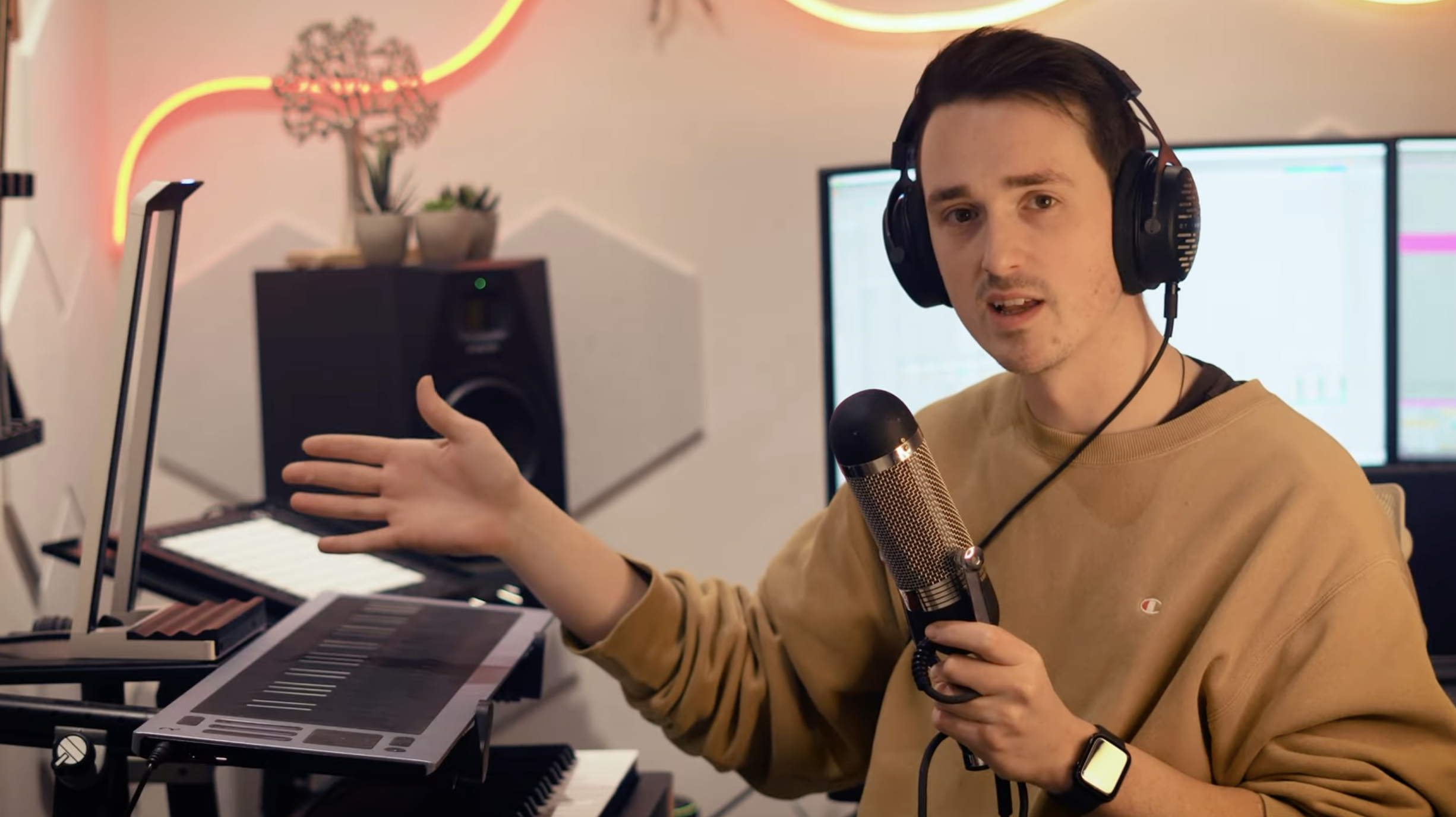 A person wearing headphones and holding a microphone gestures toward a ROLI Seaboard in a home music studio, with the instrument placed on a desk alongside a ROLI Lightpad and other audio equipment.