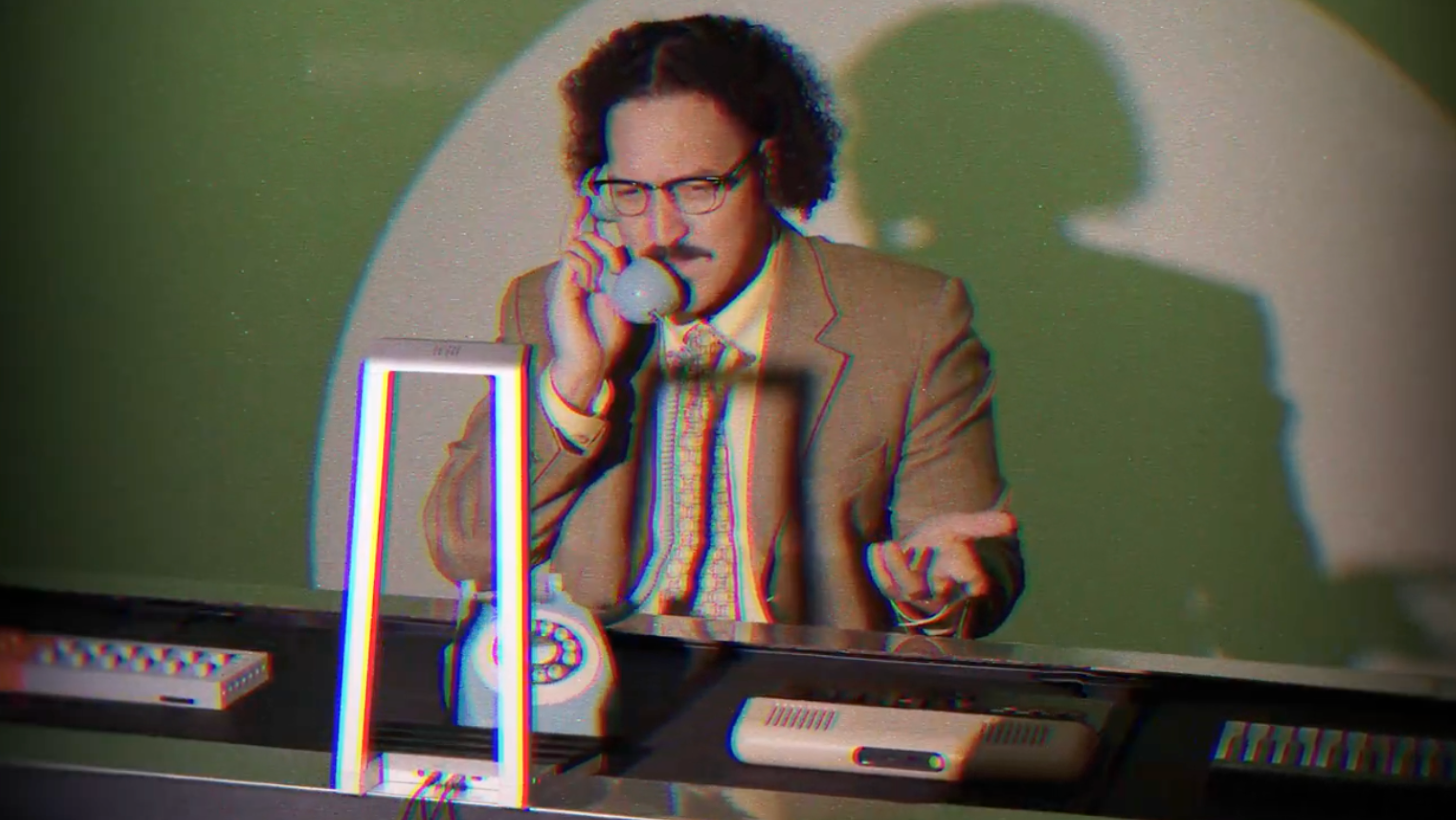 A man in a tan suit and glasses sits at a desk, speaking on a rotary phone, with a ROLI Lightpad and a ROLI Seaboard placed on the desk in a retro-styled office setting.