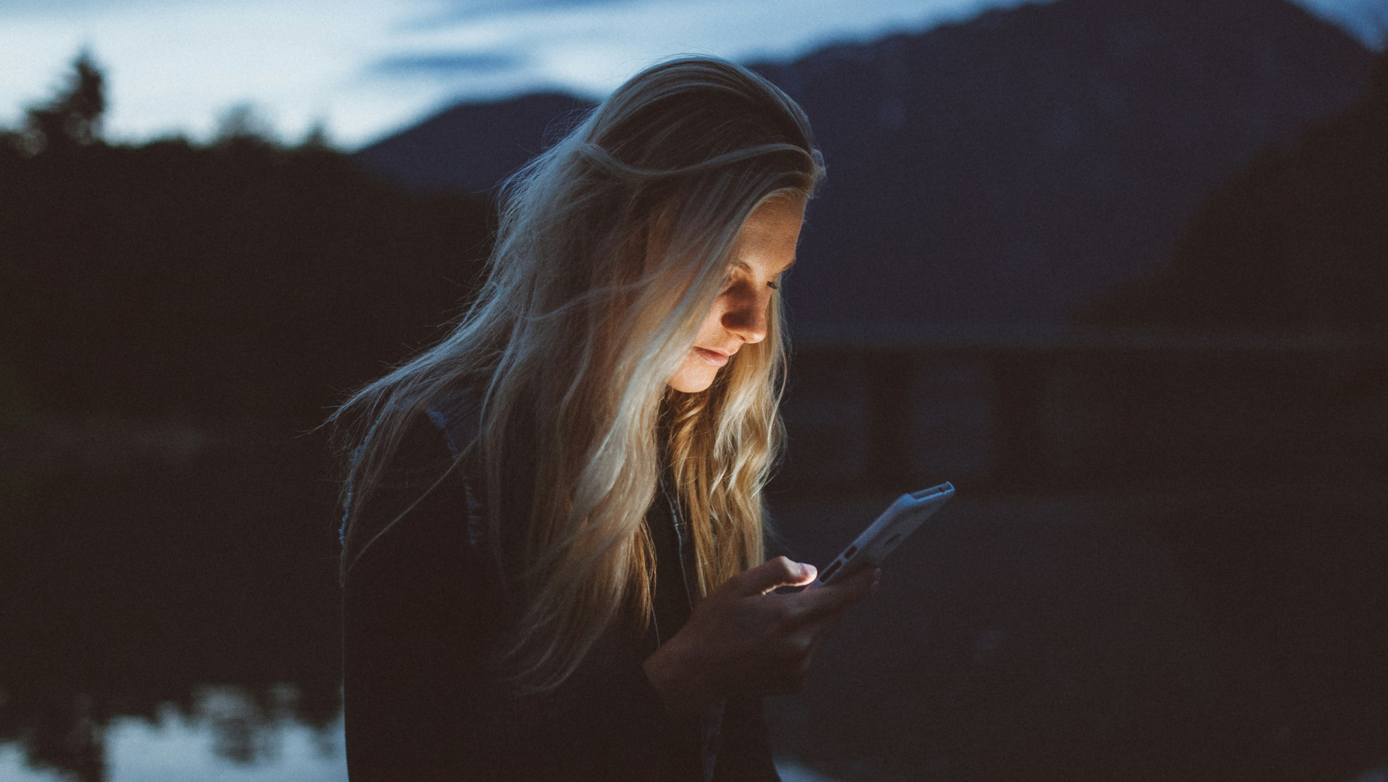 A person with long blonde hair is illuminated by the light from a smartphone, standing outdoors near a body of water at dusk with mountains in the background.