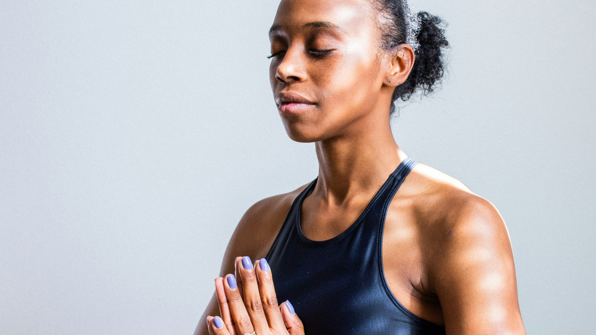 A woman in a black athletic top stands with her eyes closed and hands pressed together in a prayer position, appearing calm and focused against a plain light background.