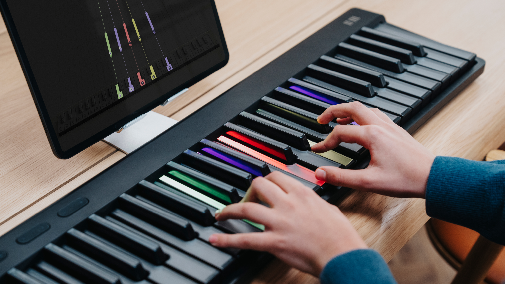 Hands playing a ROLI Piano on a wooden desk, with colorful illuminated keys and a computer screen displaying a music learning interface.