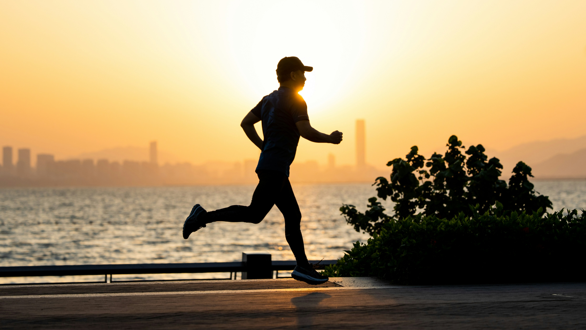 A person jogging along a waterfront path at sunset, with city buildings and mountains visible in the background.