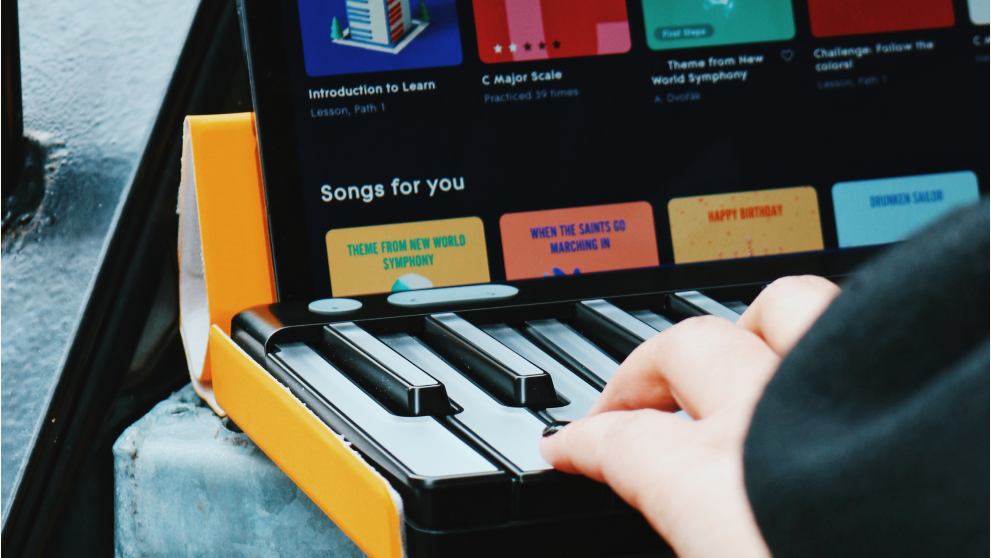 A person's hand playing a ROLI Piano on a desk, with a tablet displaying the ROLI Learn app and song selections in the background.
