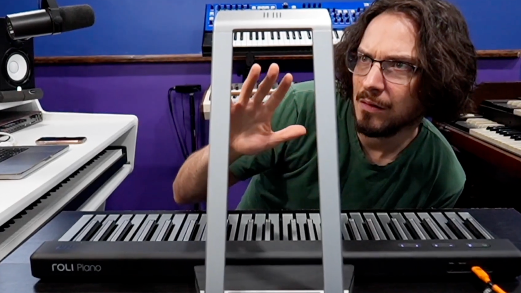 A person in a green shirt interacts with a ROLI Piano on a desk, raising their hand above the instrument, with studio equipment and keyboards visible in the background.