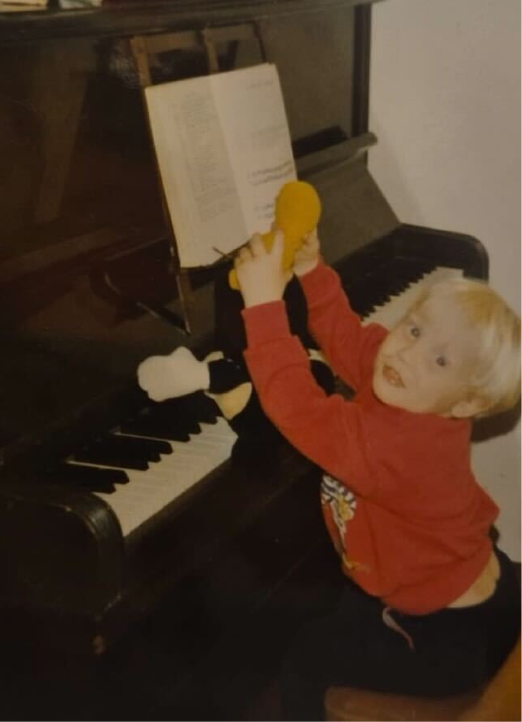 A young child in a red sweater sits at an upright piano, holding a yellow plush toy up to an open sheet music book on the music stand.