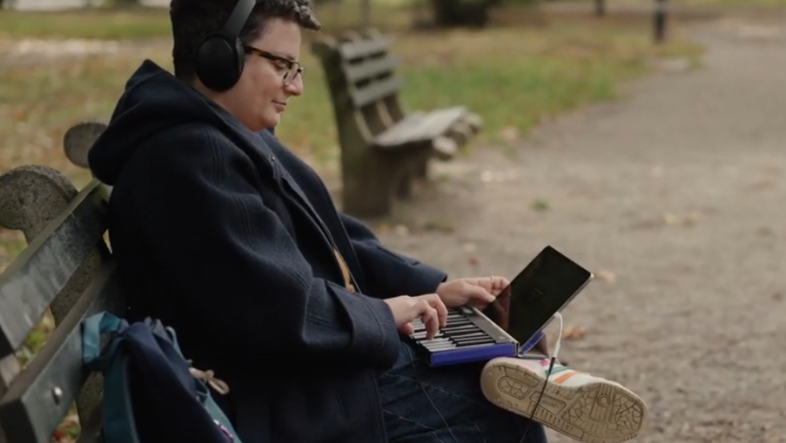 A person wearing headphones sits on a park bench, playing a ROLI Piano on their lap with a tablet propped up in front of them.