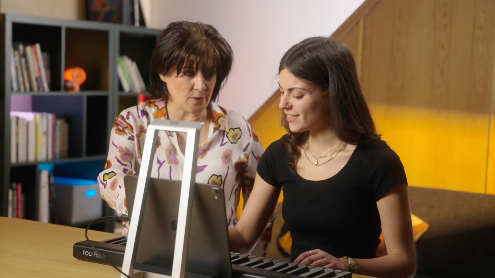 Two women sit at a table in a home setting, with one playing a ROLI Piano keyboard connected to a tablet, while the other observes and provides guidance.