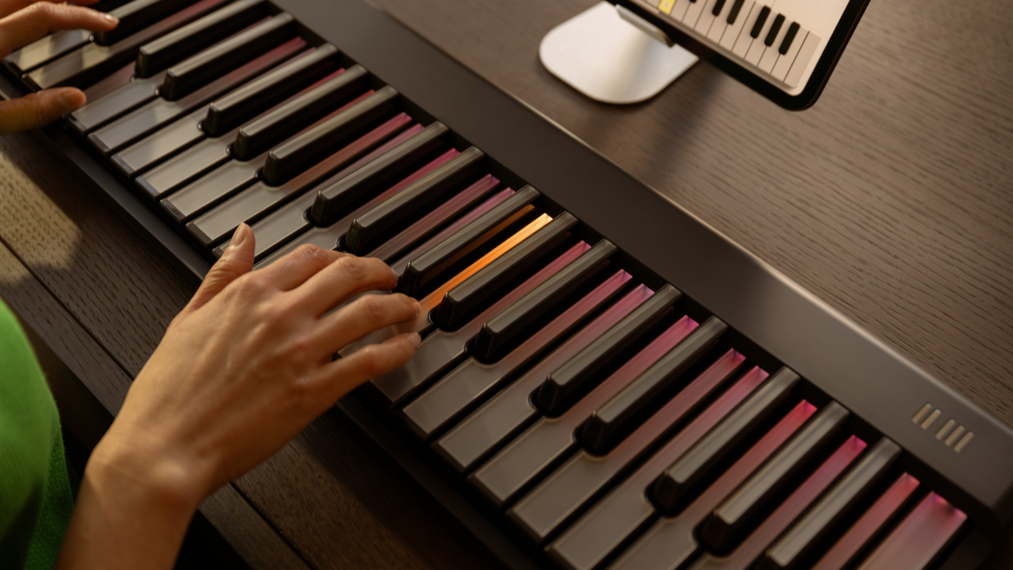 Hands playing a ROLI Piano on a wooden desk, with the instrument’s keys illuminated in pink and orange and a tablet displaying a virtual keyboard in the background.