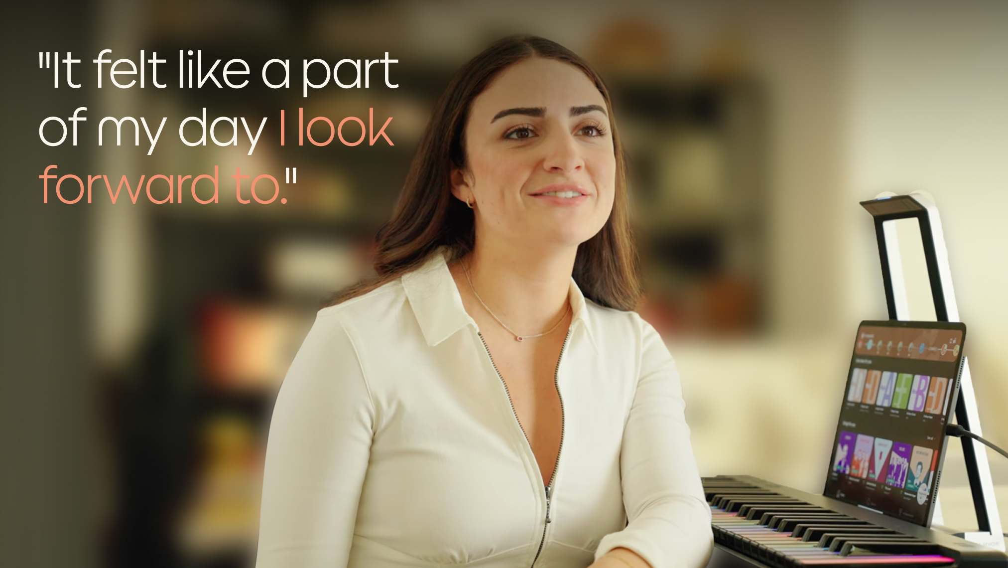 A woman sits and smiles in a well-lit room, with a ROLI Piano on a desk in front of her and a tablet displaying the ROLI Learn app propped up on the piano.