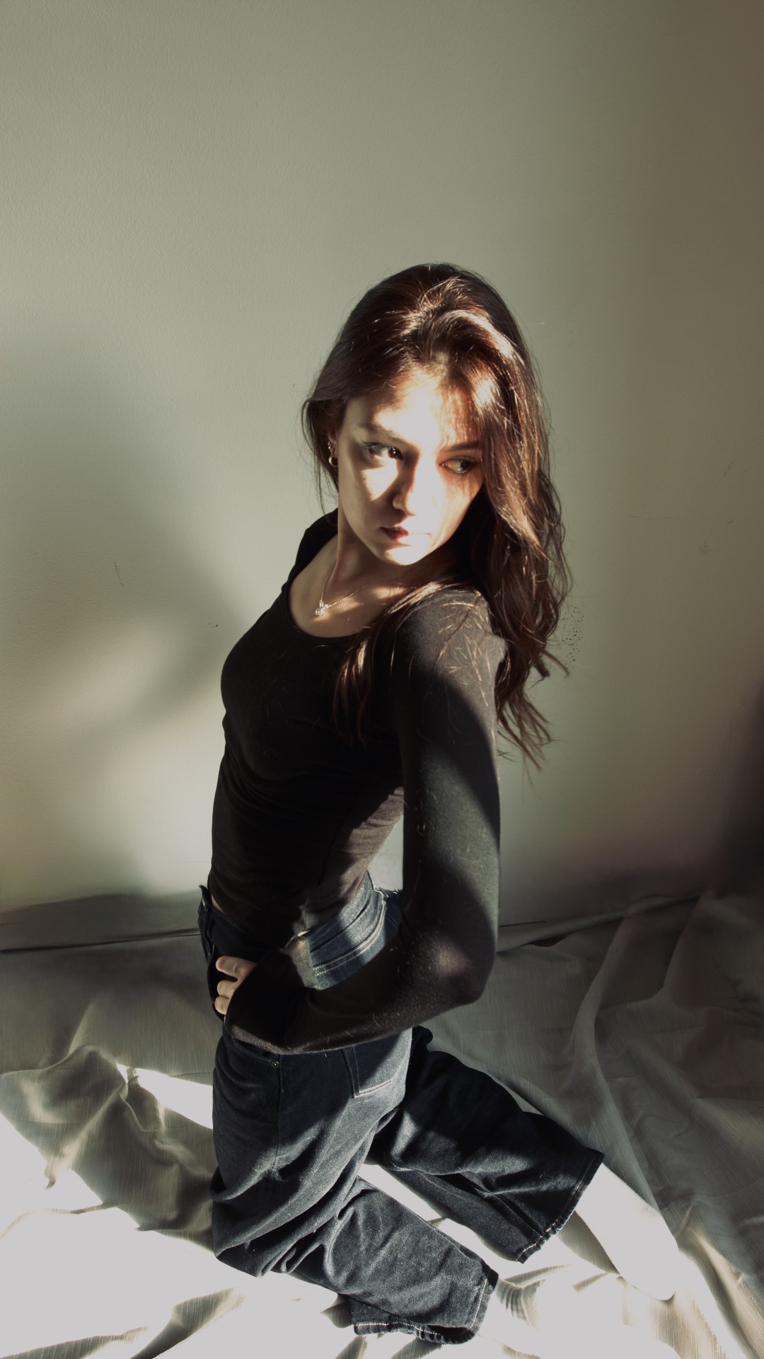 A woman with long brown hair wearing a black long-sleeve top and dark jeans kneels on a light-colored fabric, looking to the side in soft natural light against a plain wall.