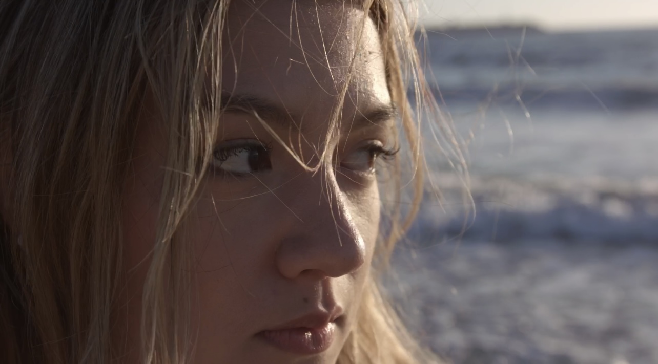 A close-up of a person's face with light brown hair, looking to the side, with strands of hair across their forehead and the ocean blurred in the background.