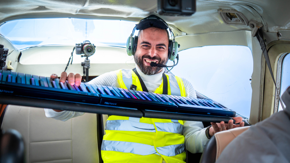A smiling man wearing a headset and high-visibility vest holds a ROLI Seaboard 2 keyboard inside a small airplane cockpit, with a camera mounted behind him.