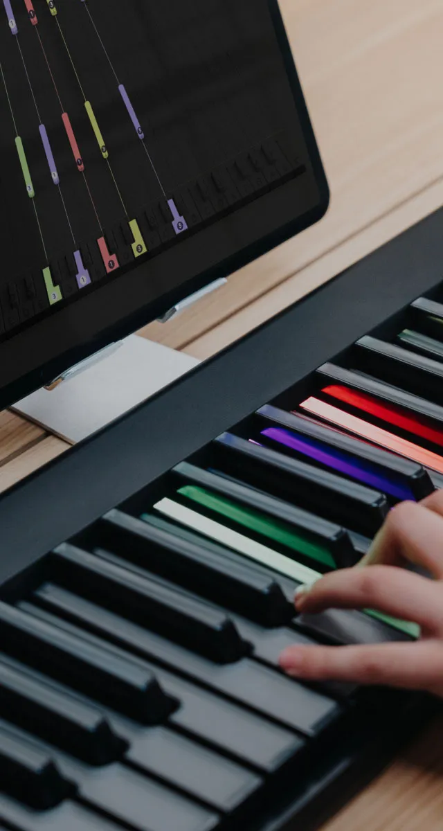 A hand playing a black ROLI Seaboard 2 keyboard with illuminated, multicolored keys, positioned on a wooden desk beneath a computer monitor displaying a music learning interface.
