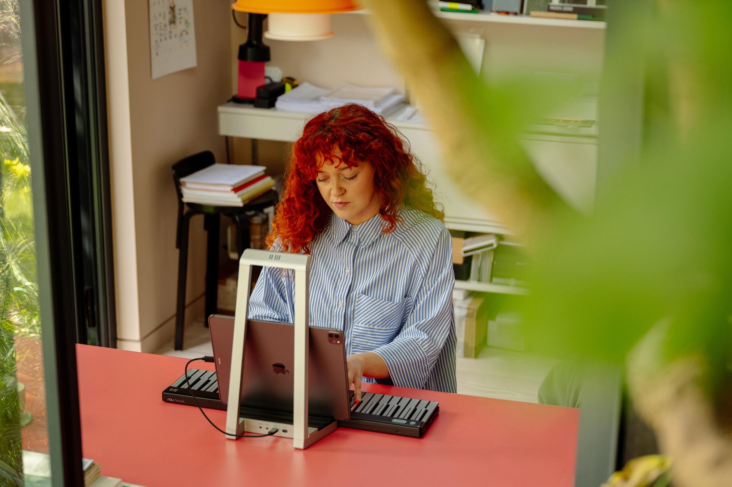 A person with curly red hair sits at a pink desk in a home office, playing a ROLI Piano with an iPad mounted in front of them.