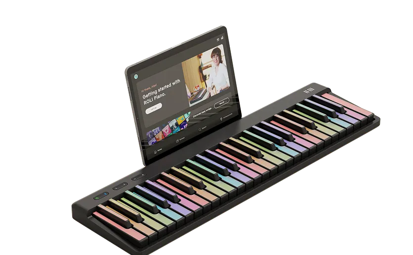 A ROLI Piano with pastel-colored keys sits on a desk, with a tablet displaying a "Getting started with ROLI Piano" lesson propped up behind the instrument.