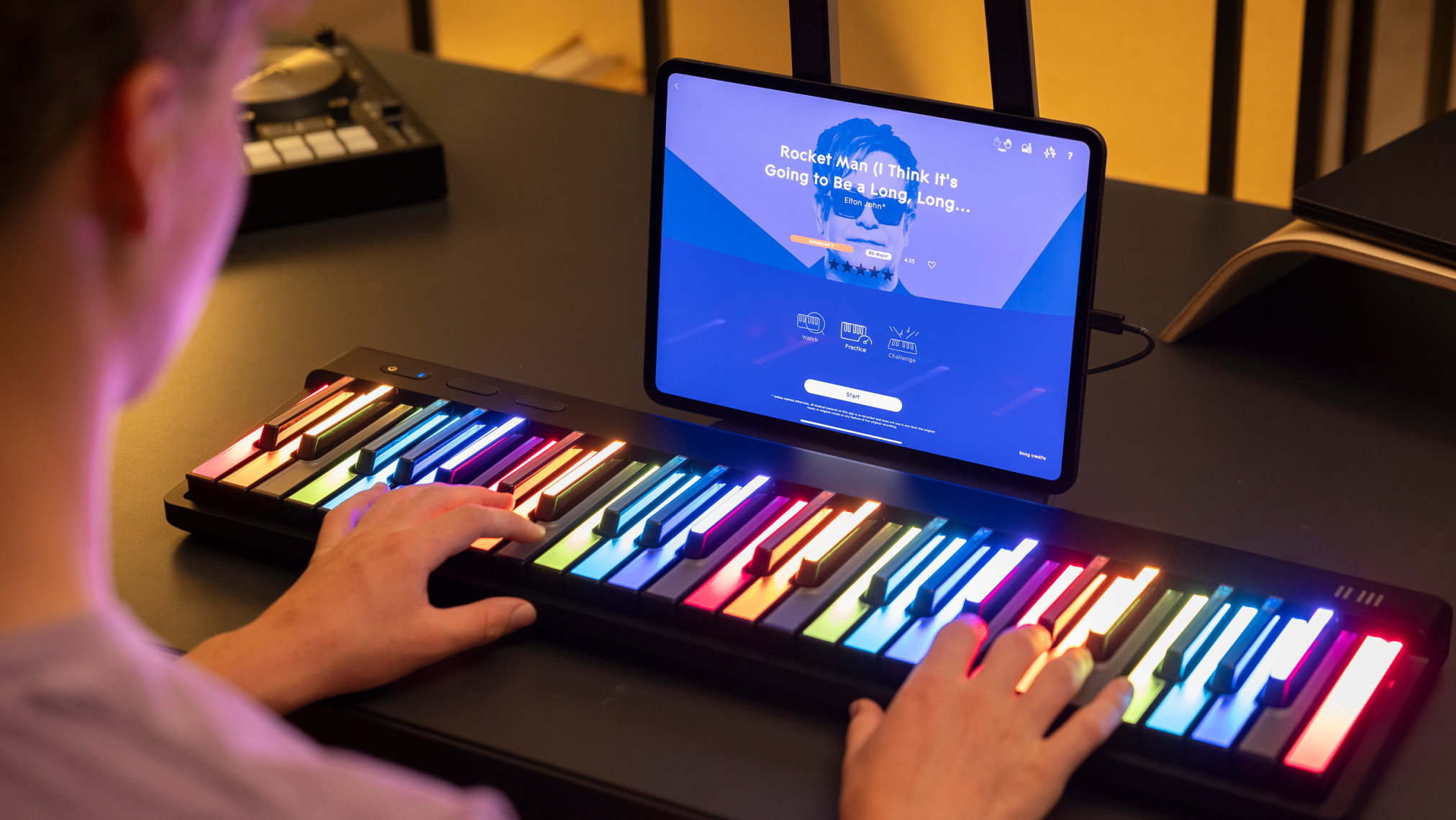 A person playing a ROLI Piano with rainbow-colored illuminated keys on a desk, while following a song tutorial for "Rocket Man" by Elton John on a connected tablet.