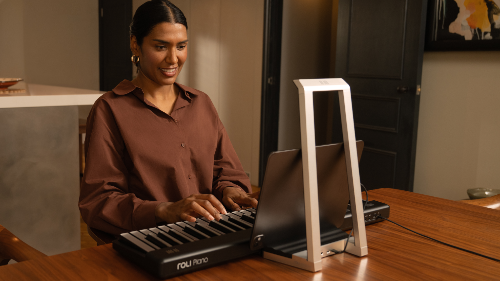 A person smiling while playing a ROLI Piano on a wooden desk, with a laptop positioned in front of the instrument in a home setting.