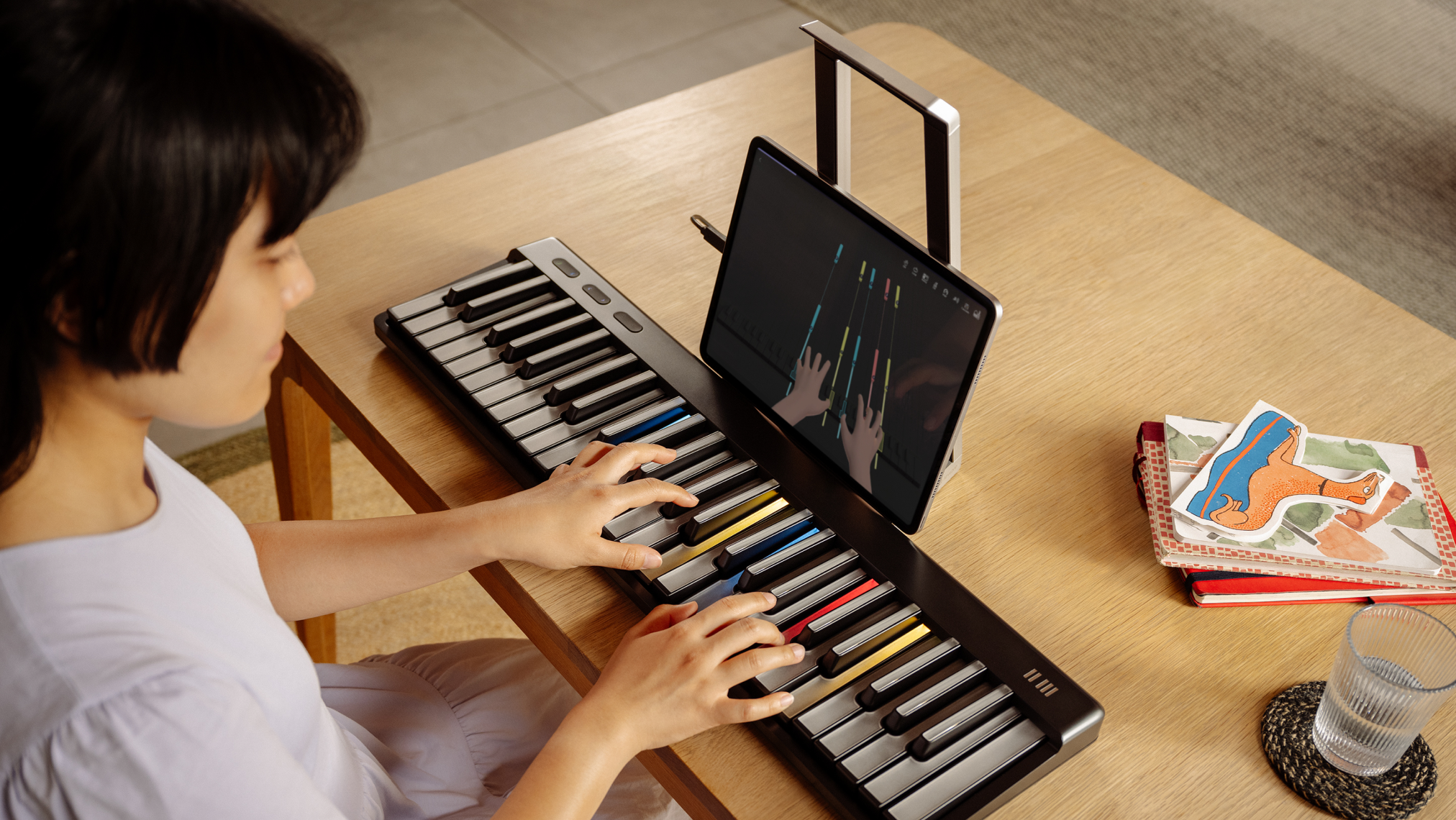 A person playing a ROLI Piano on a wooden desk, with the ROLI Learn app open on a tablet displaying a virtual keyboard and hand positions.