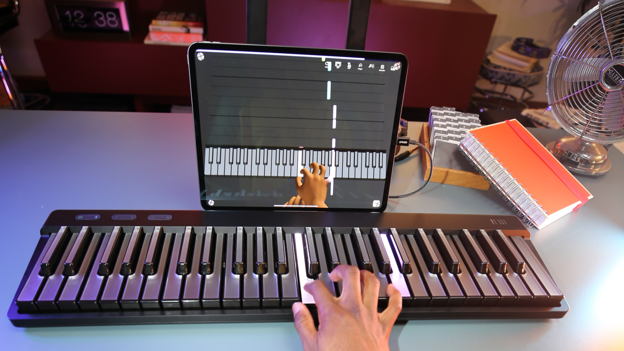 A person's hand playing a black ROLI Piano on a desk, with a tablet displaying a virtual keyboard and hand animation for learning, surrounded by a red notebook, books, and a metal fan.