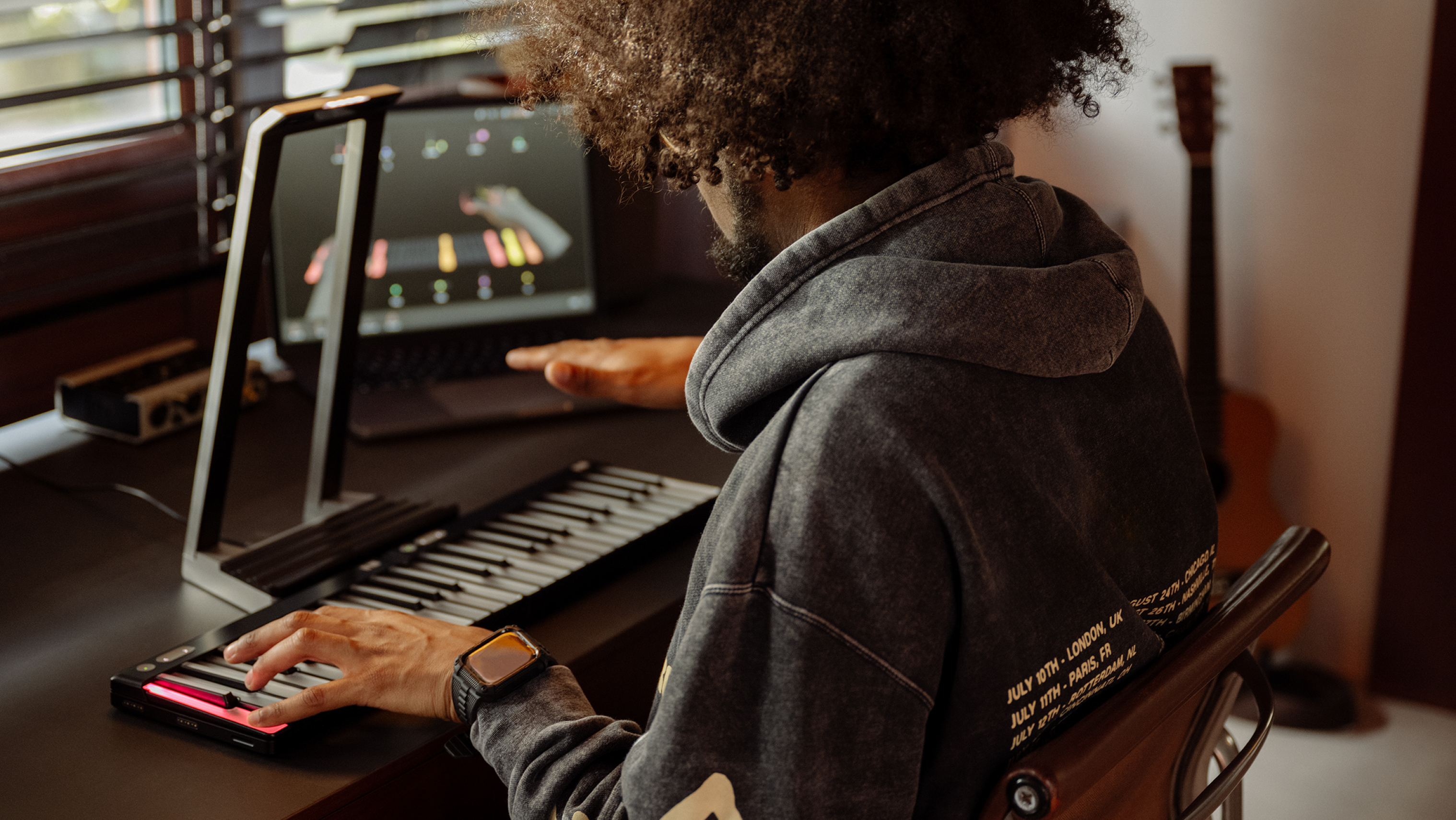 A person playing a black ROLI Seaboard on a desk, with one hand pressing the keys and the other hand hovering above the instrument, while a laptop displays music software in the background.