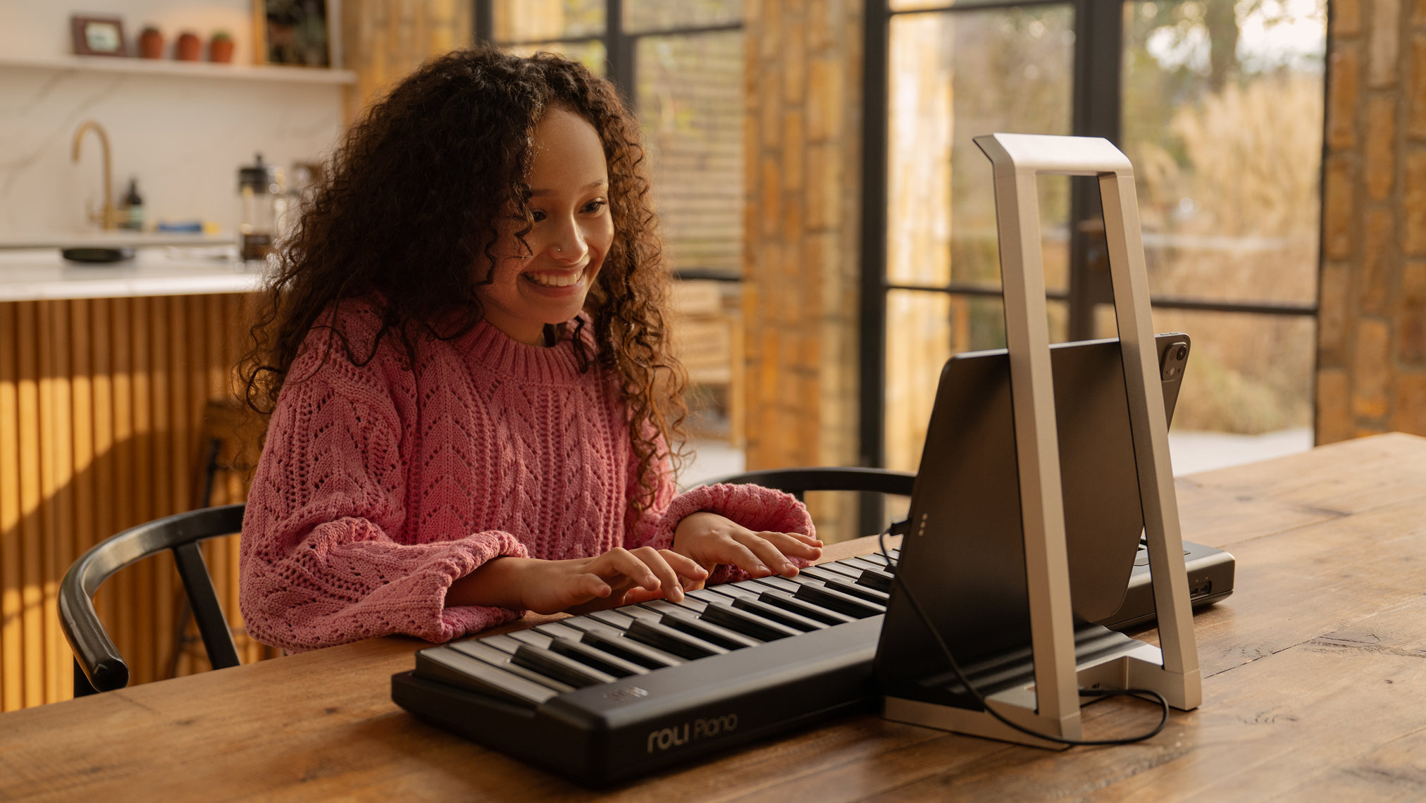 A smiling person in a pink sweater plays a ROLI Piano on a wooden table, following along with a tablet in a modern, sunlit home setting.