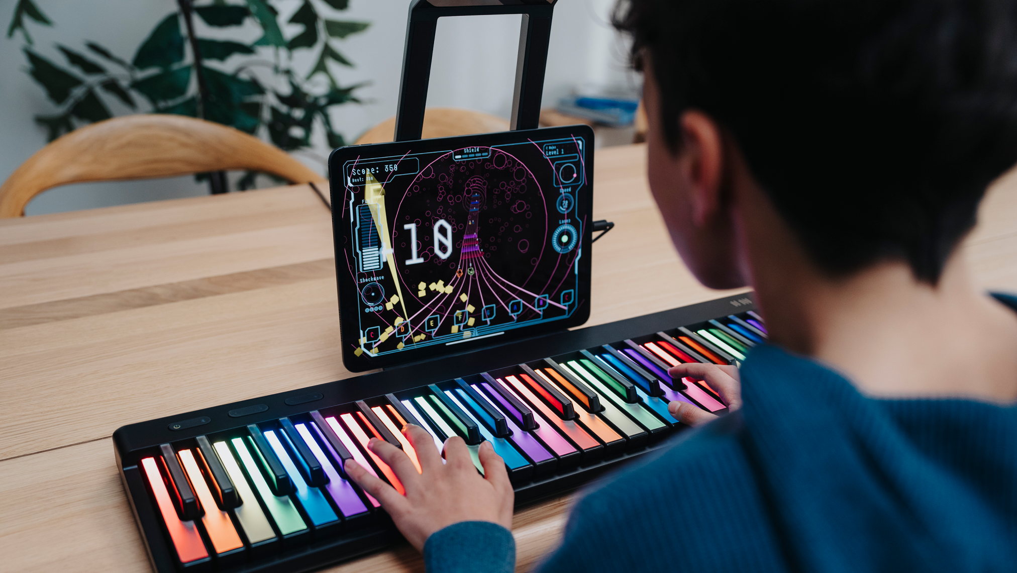 A person playing a ROLI Piano on a wooden table, with both hands on the instrument’s colorful, illuminated keys and a tablet displaying a music learning game.
