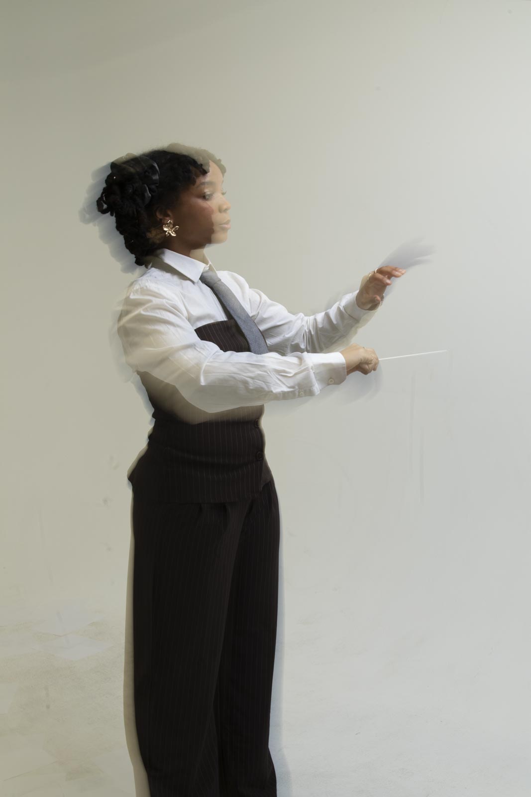 A person in a white shirt, dark vest, and tie stands against a plain background, with a motion blur effect showing their arms raised and hands gesturing forward.