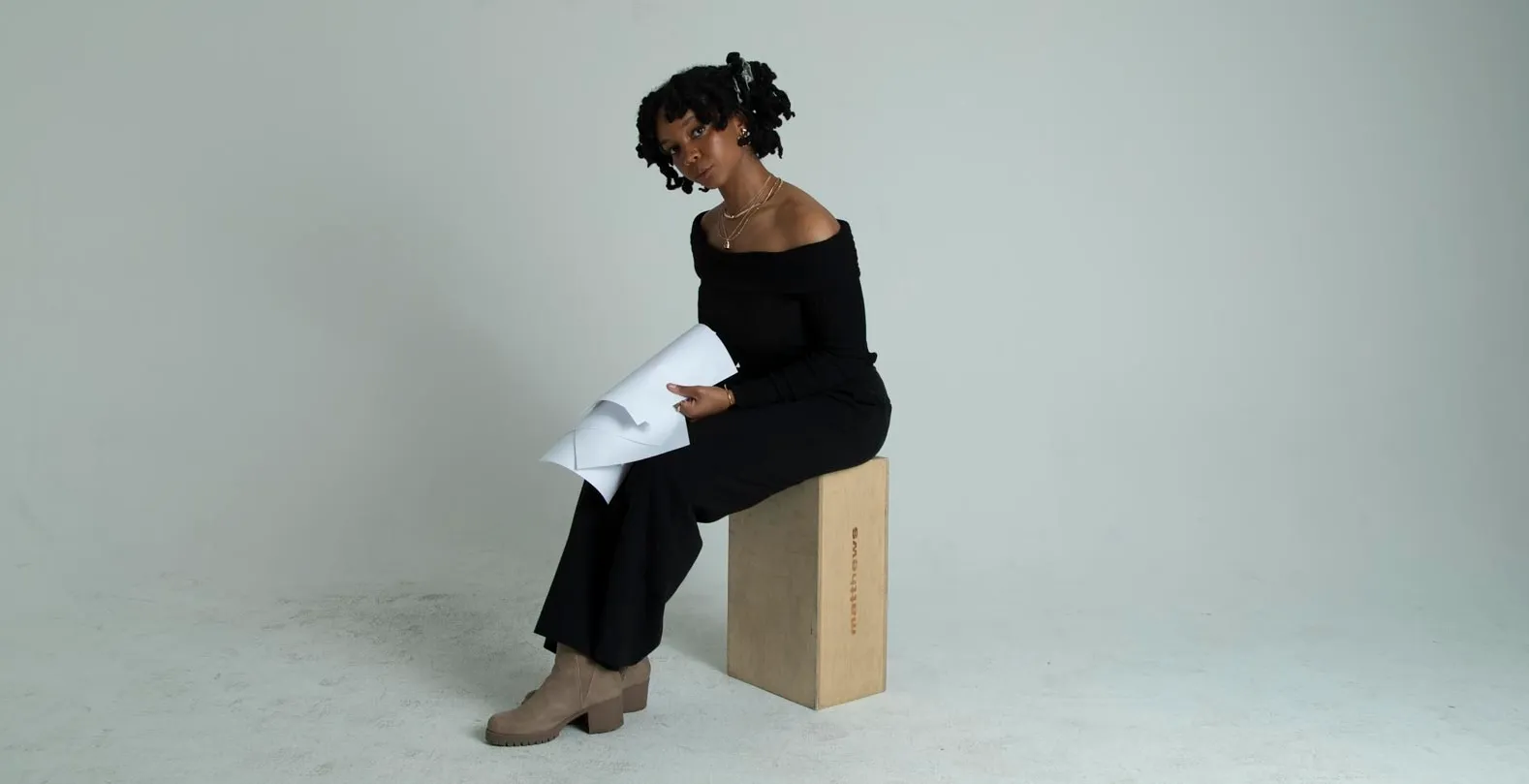 A person wearing a black off-the-shoulder top and black pants sits on a wooden box in a minimalist studio, holding rolled-up white papers and looking toward the camera.