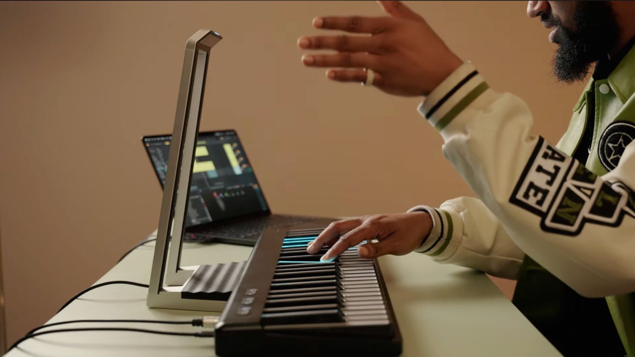 A person playing a black ROLI Seaboard 2 on a light-colored desk, with one hand on the keys and a laptop displaying music software in the background.