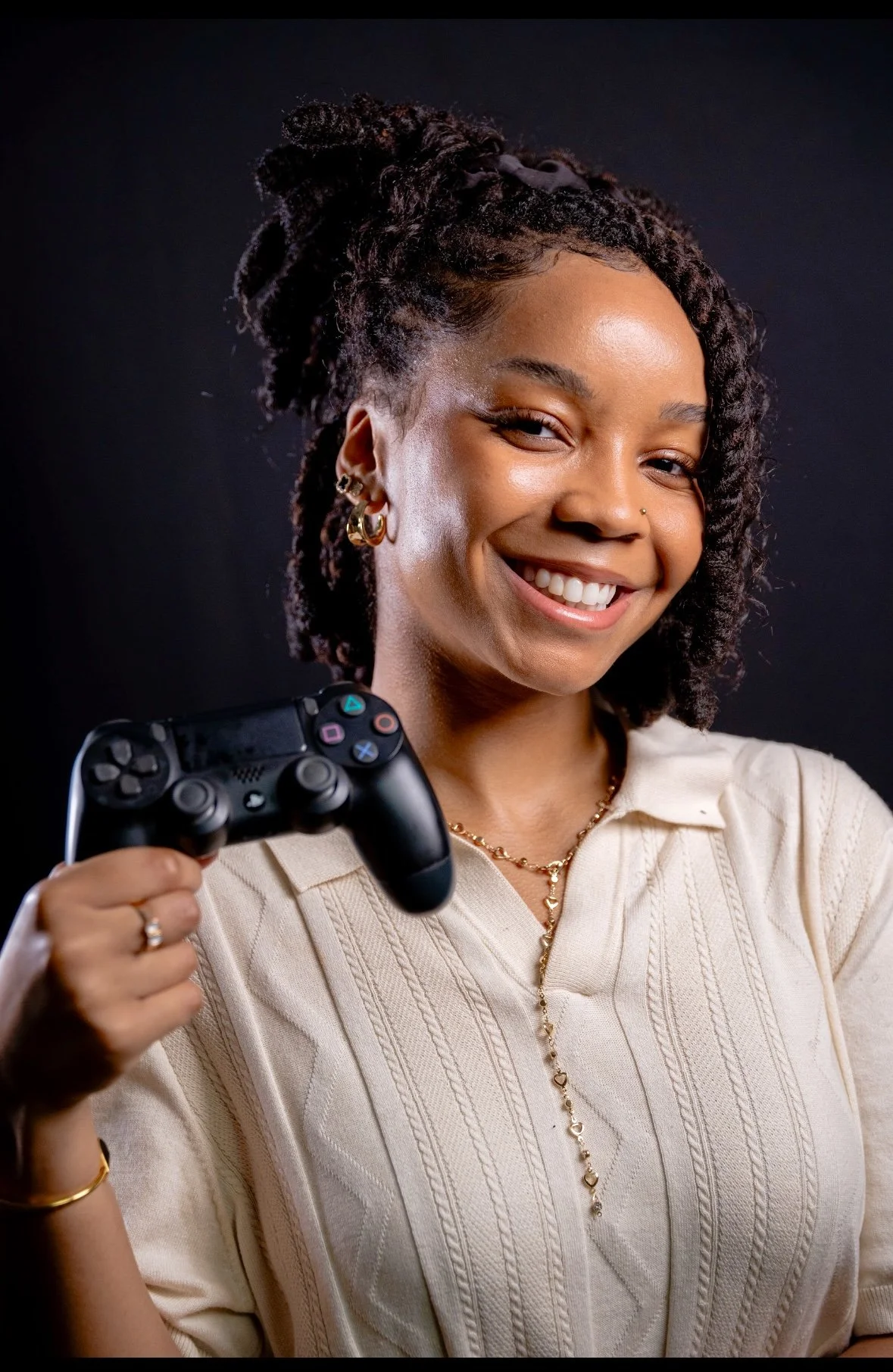 A smiling person wearing a cream-colored shirt holds a black video game controller in their right hand against a dark background.