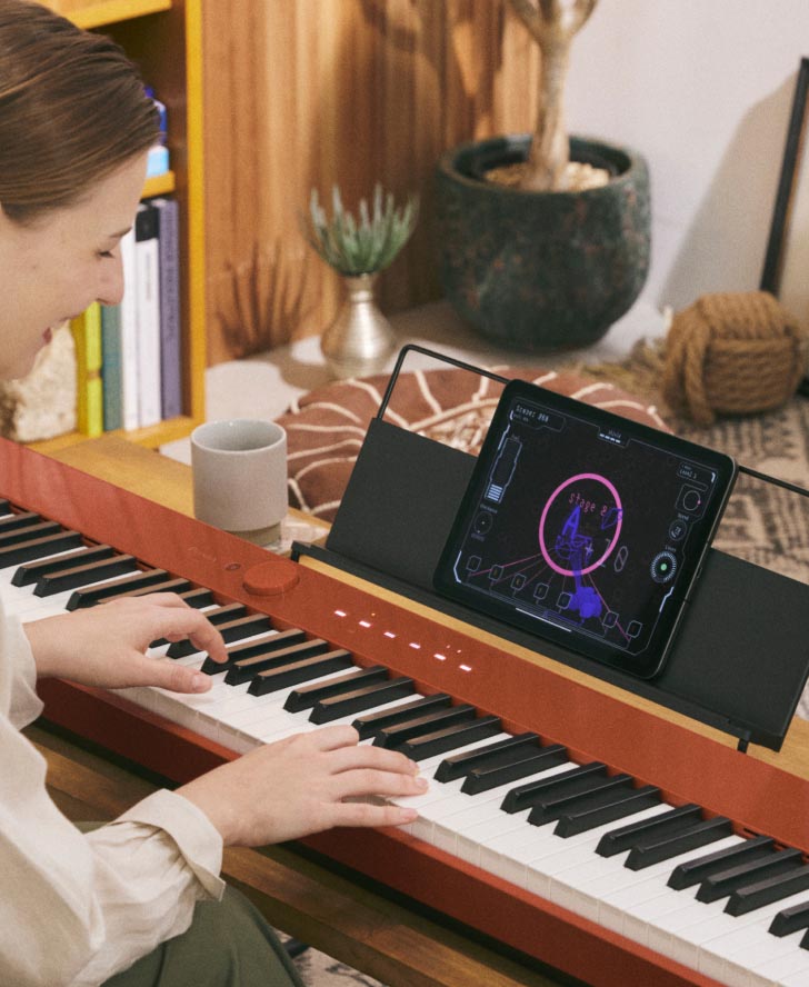 A person playing a red ROLI Piano on a wooden desk in a cozy home setting, with a tablet displaying a music app placed on the piano's built-in stand.
