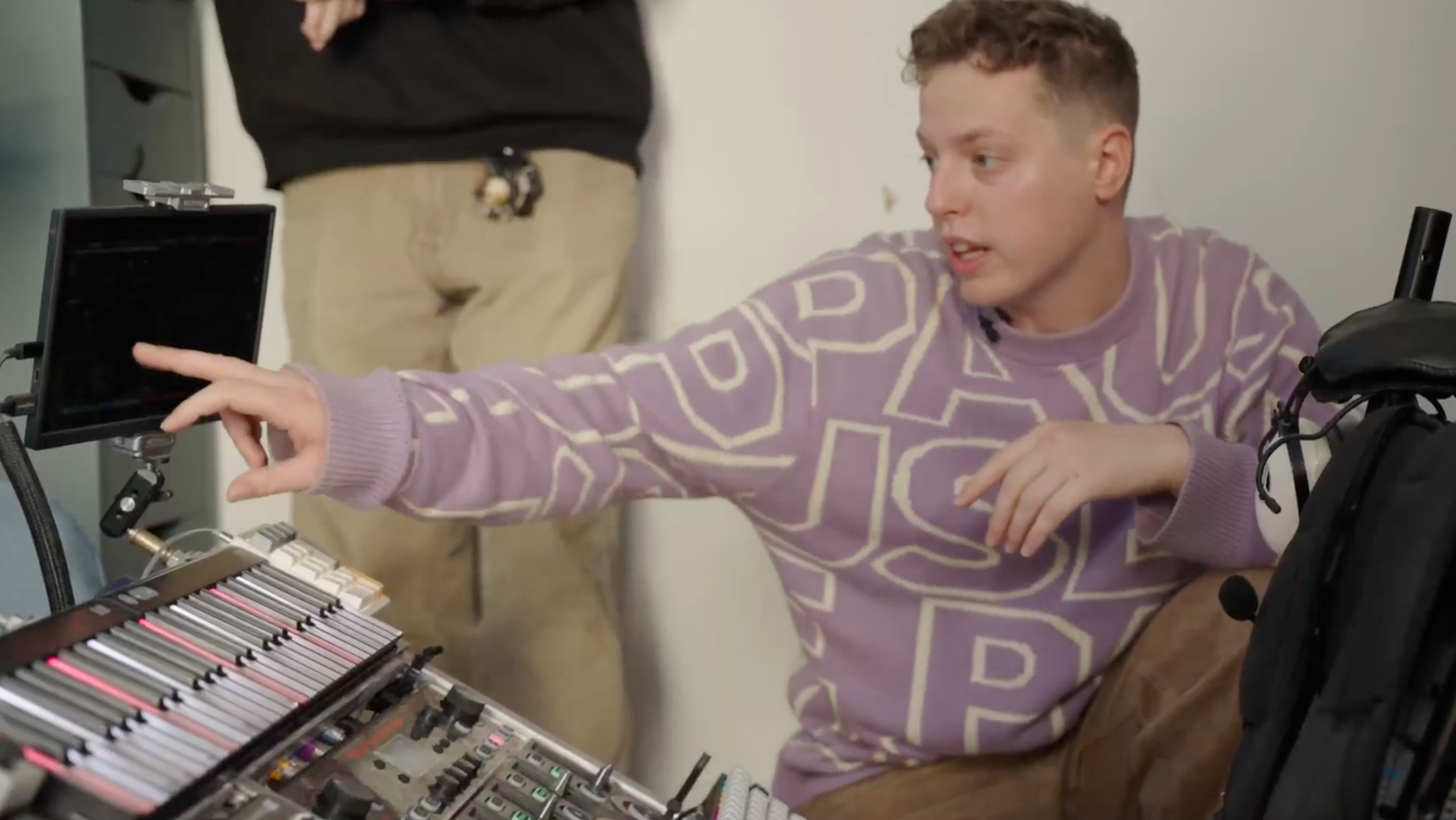 A person in a purple patterned sweater points at a monitor while sitting beside an electronic music setup with multiple control surfaces and faders in a studio environment.
