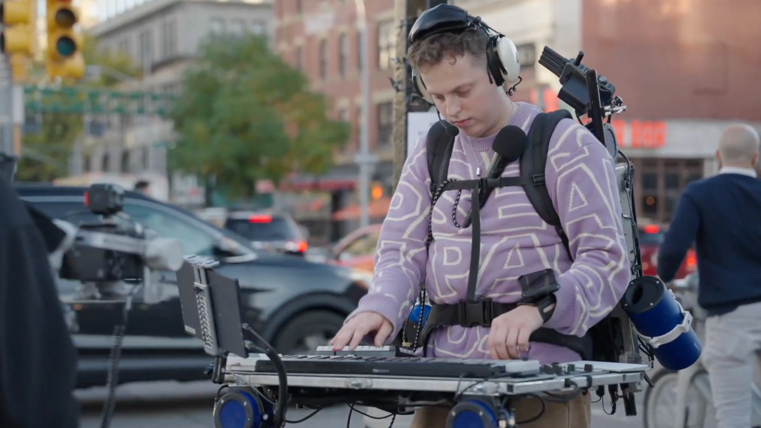 A person wearing headphones and a lavender patterned sweater plays a portable keyboard synthesizer mounted on a wearable rig in an urban outdoor setting, with a microphone attached to their chest and various electronic equipment strapped to their backpack.