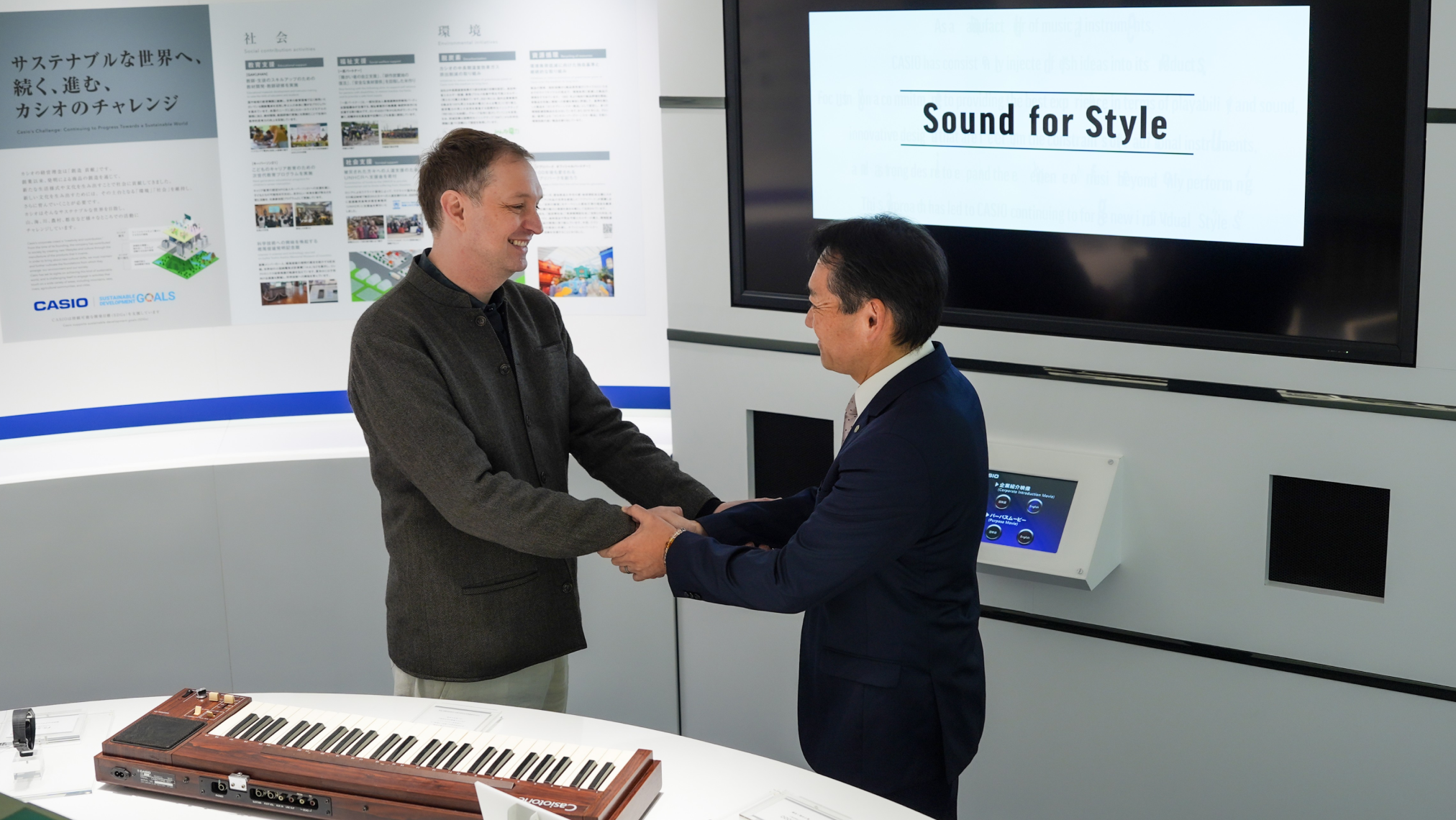 Two men shaking hands in a modern showroom, with a wooden electronic keyboard on the counter in front of them and a large screen behind displaying the text "Sound for Style."