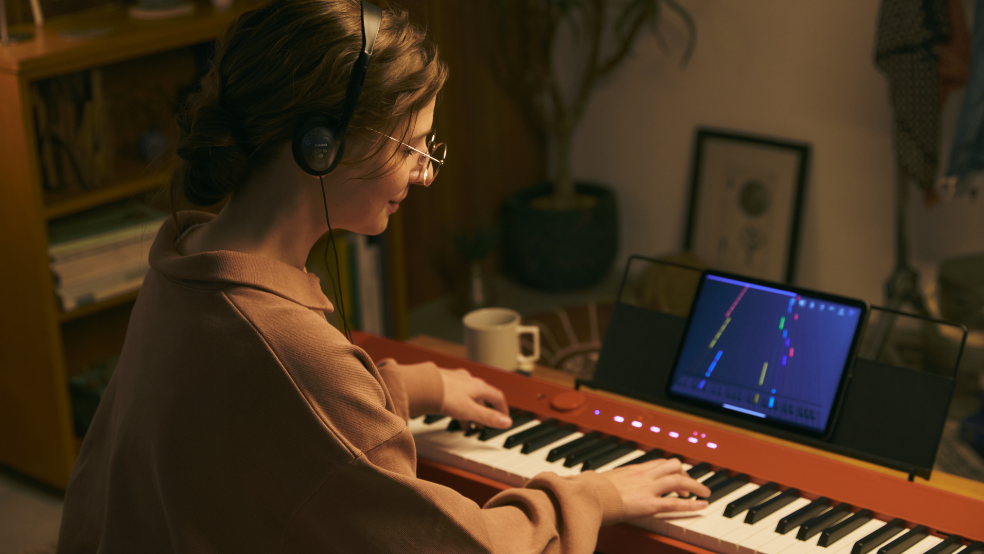 A person wearing headphones plays a ROLI Piano on a wooden desk, with a tablet displaying the ROLI Learn app above the keyboard in a cozy home setting.