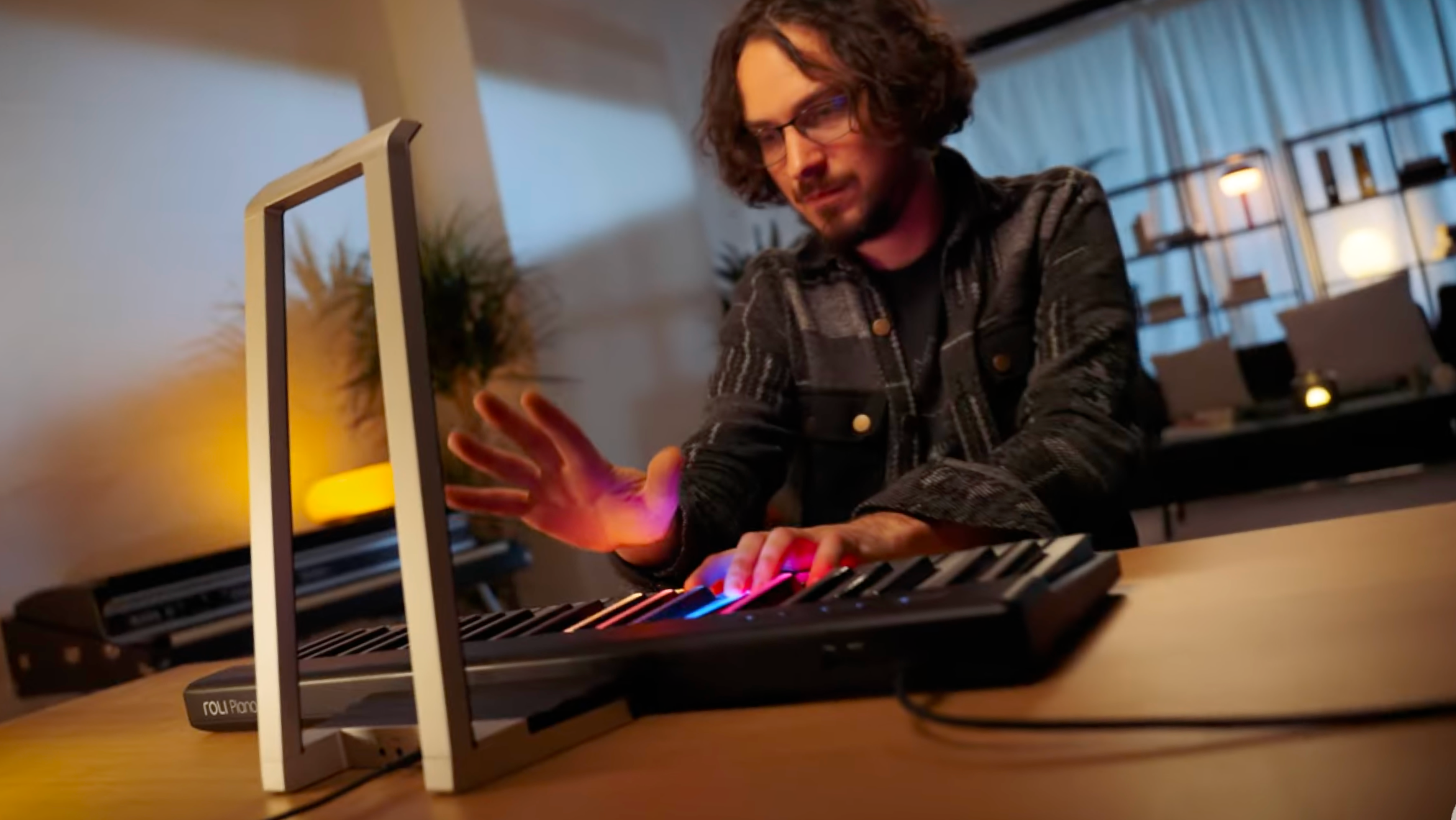 A person playing a ROLI Piano on a wooden desk in a warmly lit room, with one hand pressing illuminated keys and the other hand hovering above the keyboard.