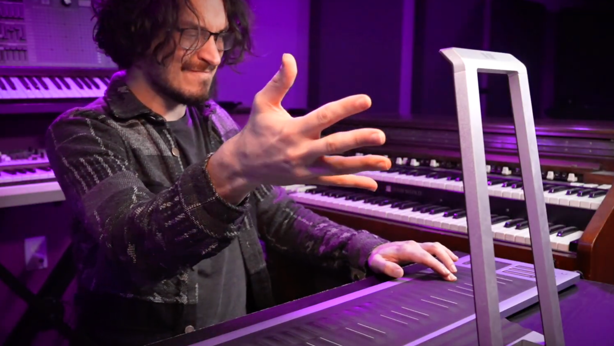 A person in a studio setting interacts expressively with a ROLI Seaboard, with one hand playing the instrument's black silicone surface and the other hand raised in a gestural motion; multiple keyboards are visible in the background under purple lighting.