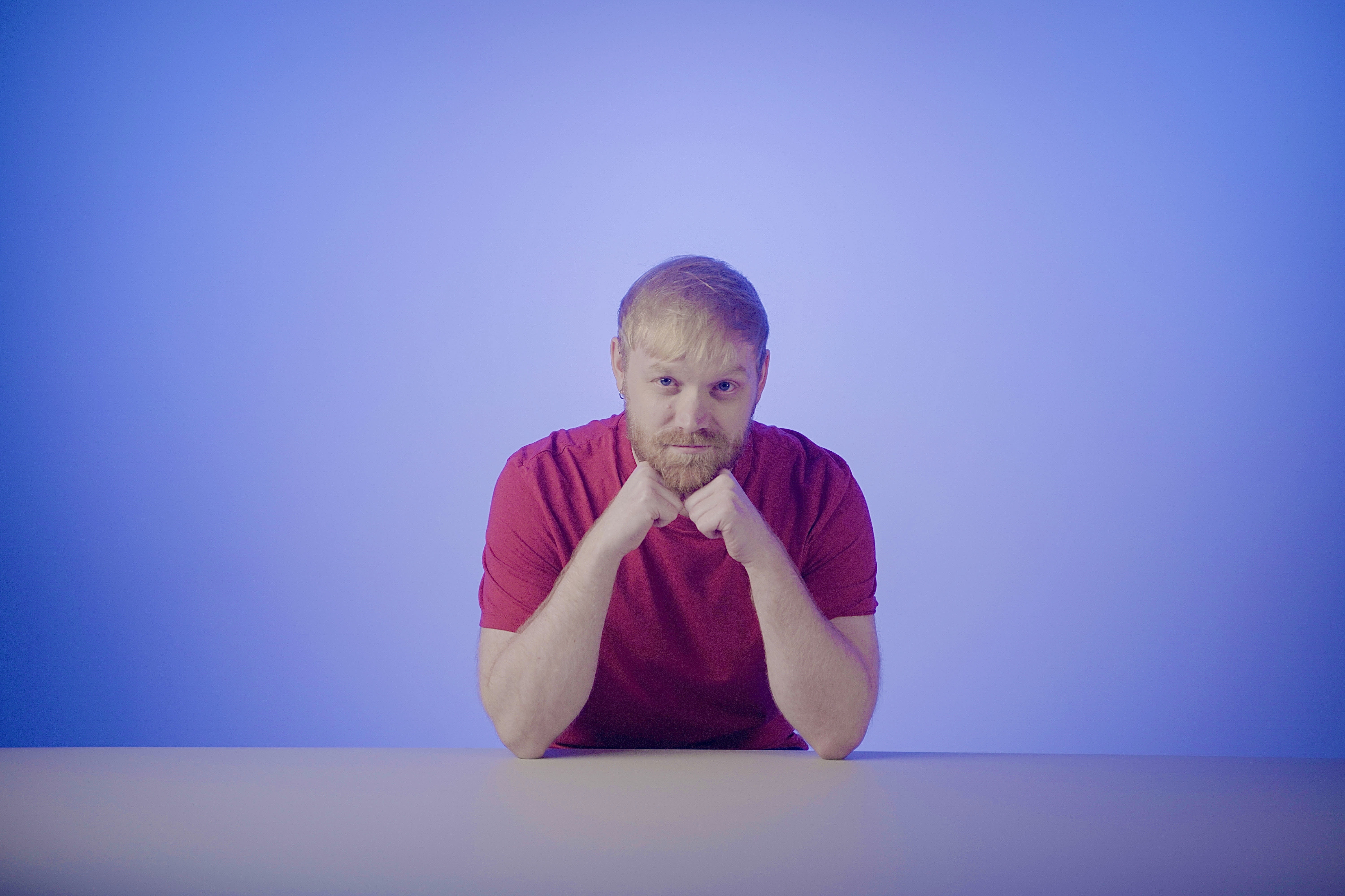 A man with blond hair and a beard sits at a table, resting his chin on his fists, against a blue gradient background.