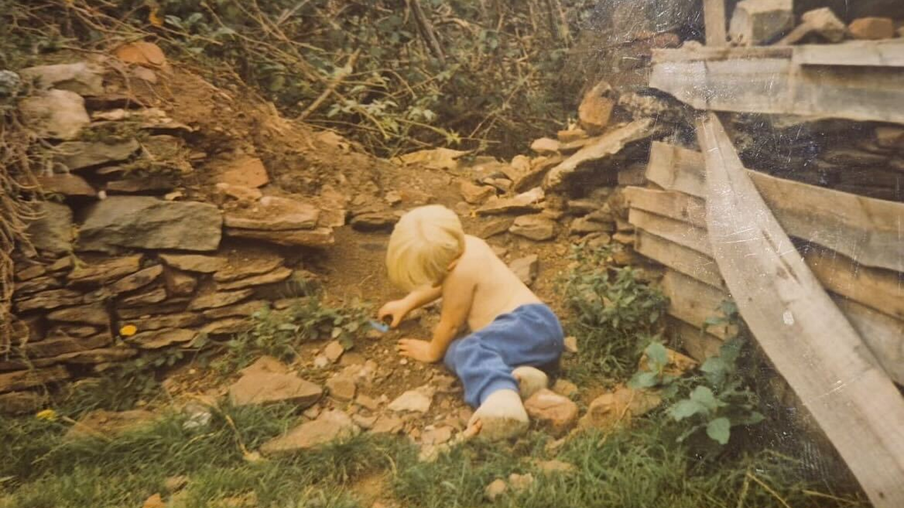 A young child with blonde hair, shirtless and wearing blue pants, is playing in the dirt next to a rustic stone wall and wooden planks in an outdoor, overgrown garden area.