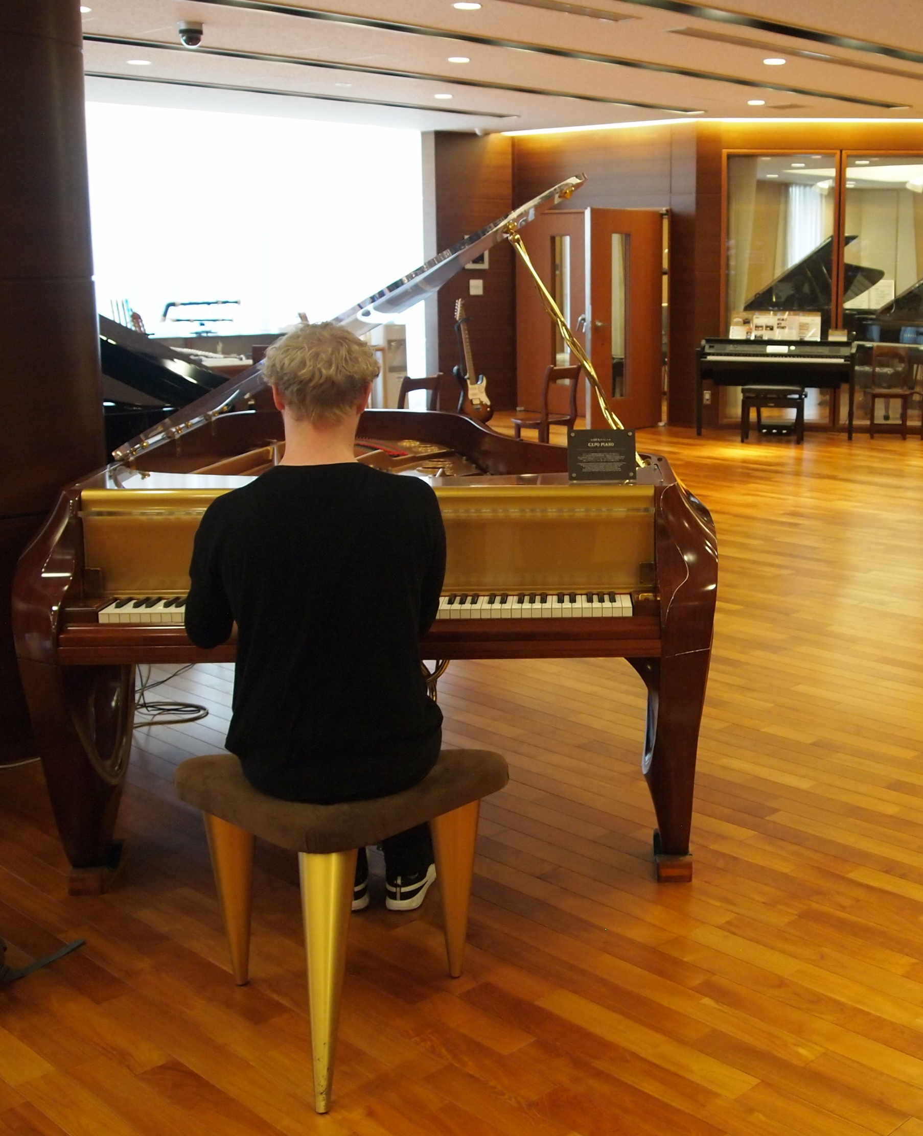A person with short blond hair sits on a brown bench playing a grand piano in a spacious showroom with wooden floors, surrounded by other pianos and musical instruments.