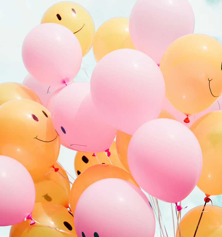 A cluster of yellow and pink balloons with smiley faces drawn on them, floating against a bright blue sky.