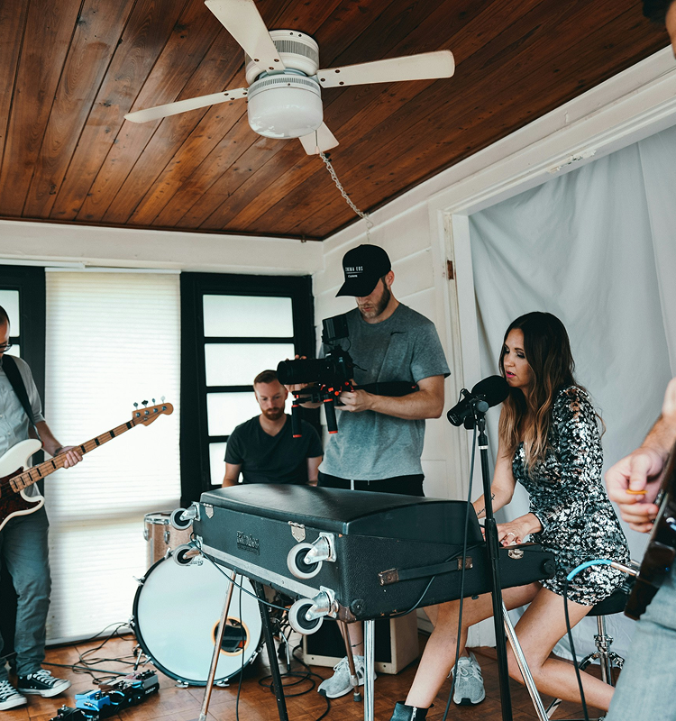 A woman in a sequined dress plays a black electric piano and sings into a microphone, accompanied by a band with a bassist, drummer, and a videographer filming the performance in a cozy, wood-paneled room.
