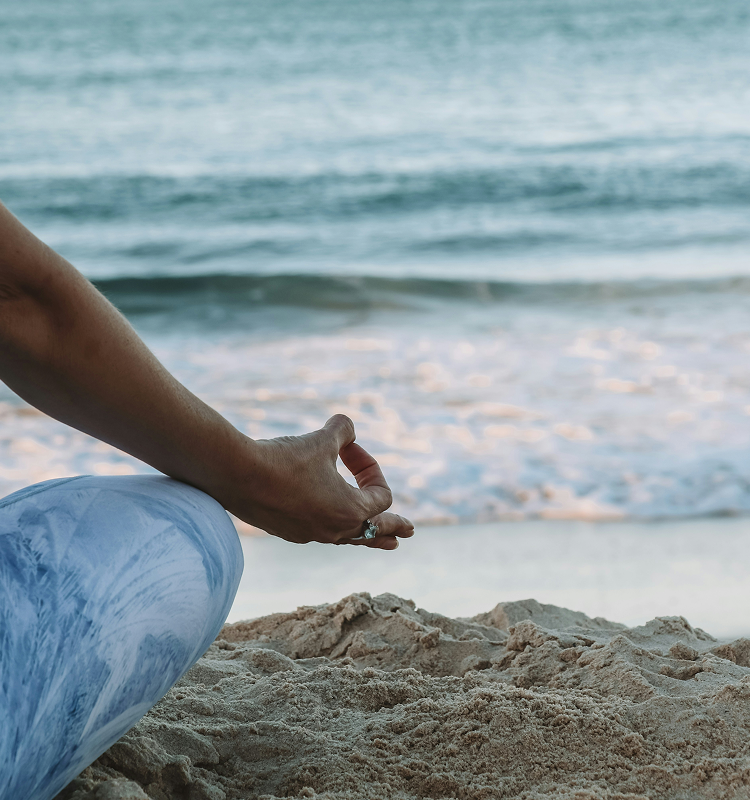 A person sitting cross-legged on sandy beach with one hand resting on their knee in a meditation pose, facing the ocean waves.