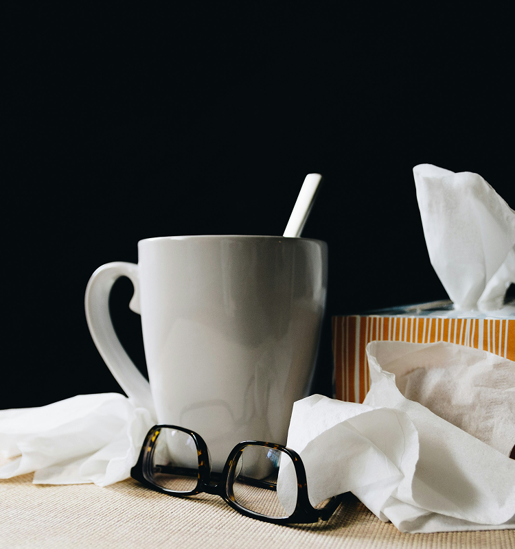 A white mug with a spoon inside sits on a table next to a box of tissues and a pair of black-framed eyeglasses, with several loose tissues scattered around.