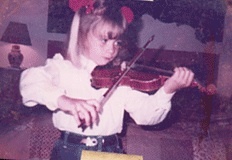 A young child with blonde hair and glasses plays a violin in a living room, wearing a white blouse and red hair bows.