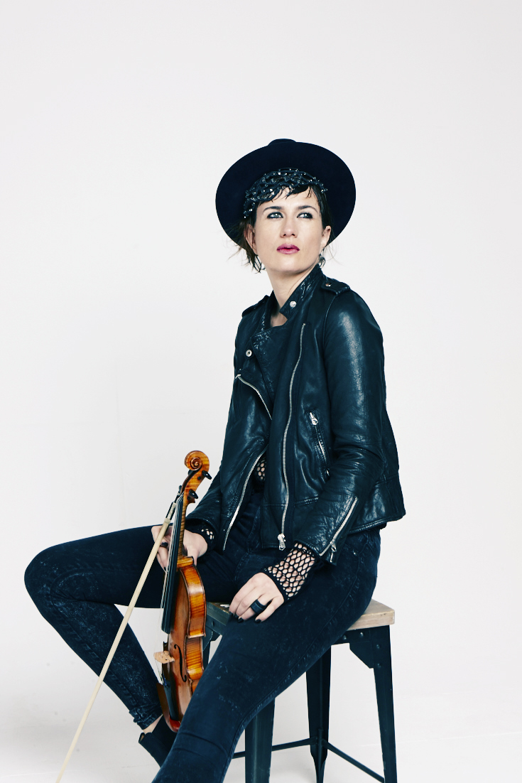 A person wearing a black leather jacket, dark pants, and a black hat sits on a stool holding a violin and bow, looking off to the side against a plain white background.
