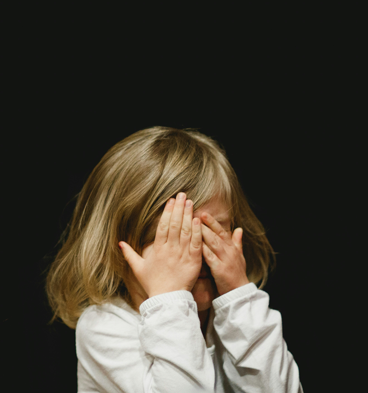 A young child with blonde hair wearing a white shirt covers their face with both hands against a black background.